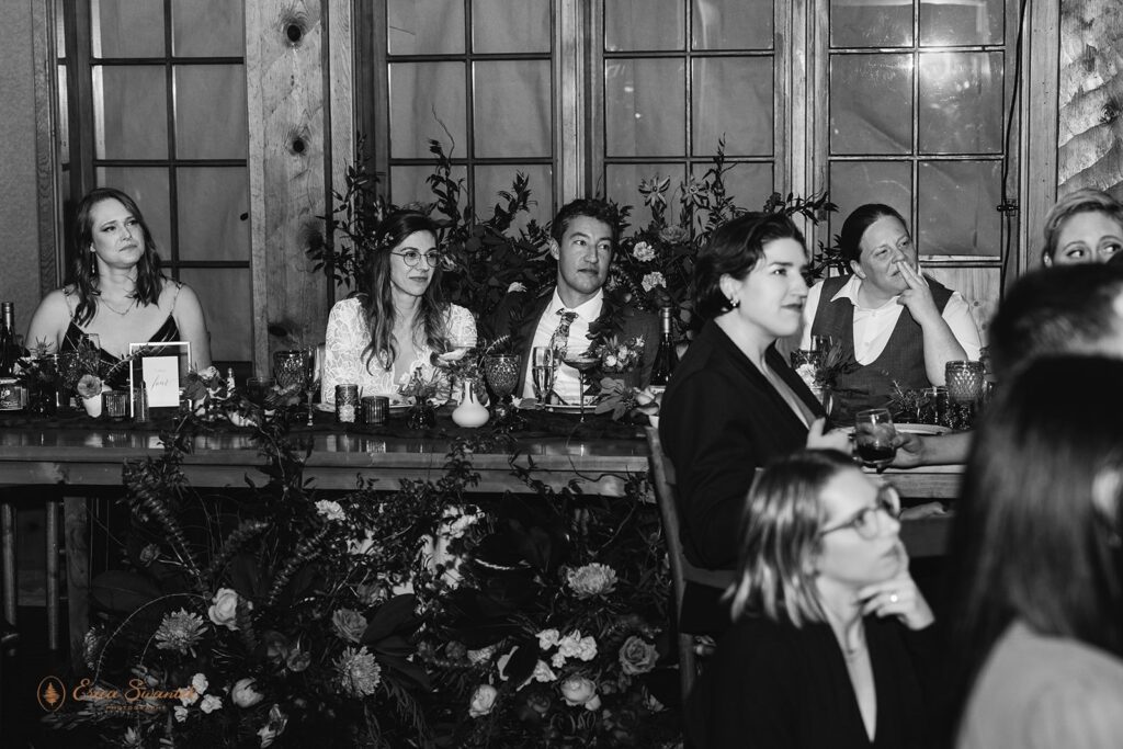 bride and groom and wedding guests sitting at the reception table, listening intently to a speech