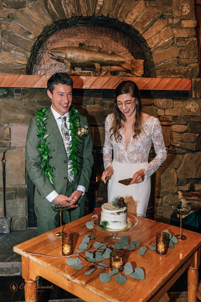 Bride and groom cut a small white cake decorated with pinecones and greenery inside a cozy cabin with a stone fireplace in the background.