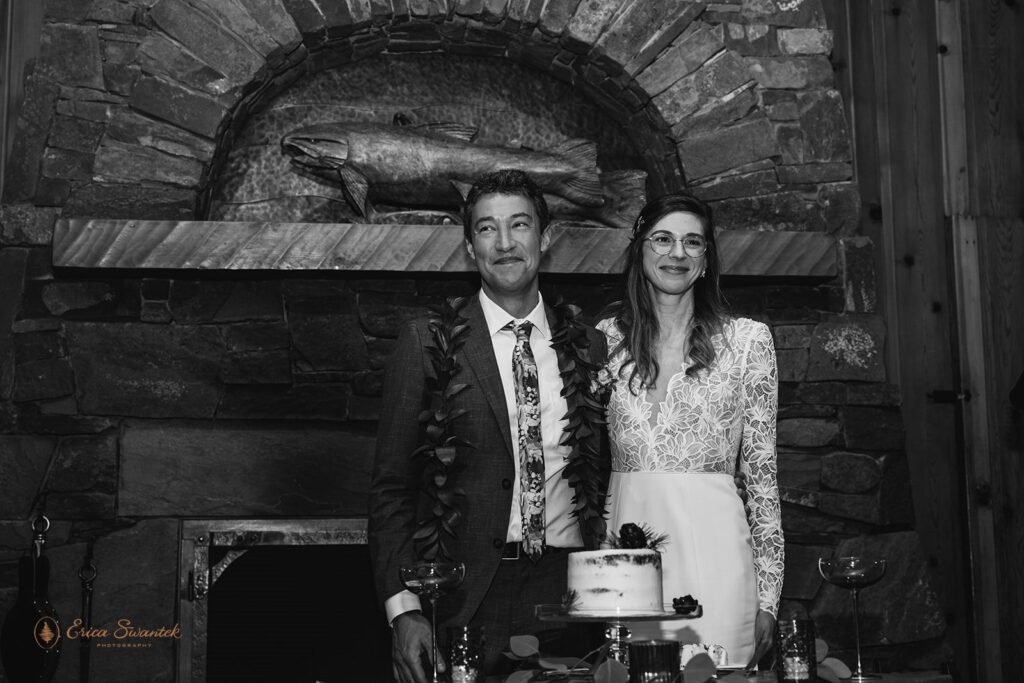 Bride and groom cut a small white cake decorated with pinecones and greenery inside a cozy cabin with a stone fireplace in the background.