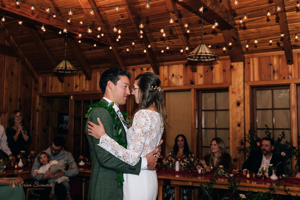 Bride and groom share their first dance inside a warmly lit wooden cabin, surrounded by friends and family who are smiling and clapping.