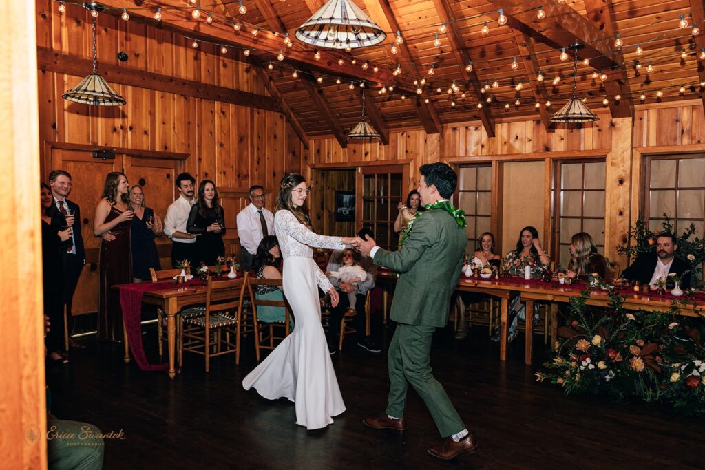 Bride and groom share their first dance inside a warmly lit wooden cabin, surrounded by friends and family who are smiling and clapping.