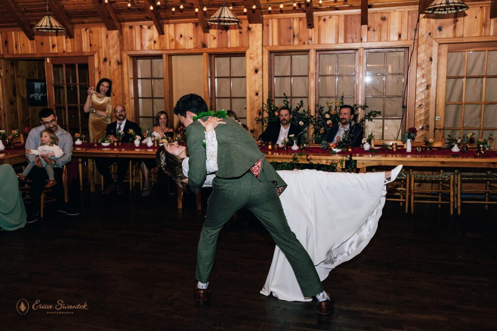 Bride and groom share their first dance inside a warmly lit wooden cabin, surrounded by friends and family who are smiling and clapping.