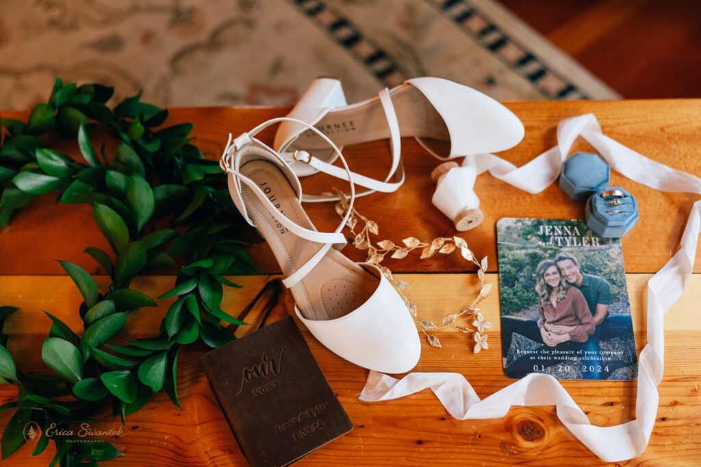 Flat lay of wedding items including a green leaf lei, white wedding shoes, a leather-bound vow book, a wedding invitation featuring a photo of Jenna and Tyler, blue ring boxes with wedding rings, and a white ribbon.