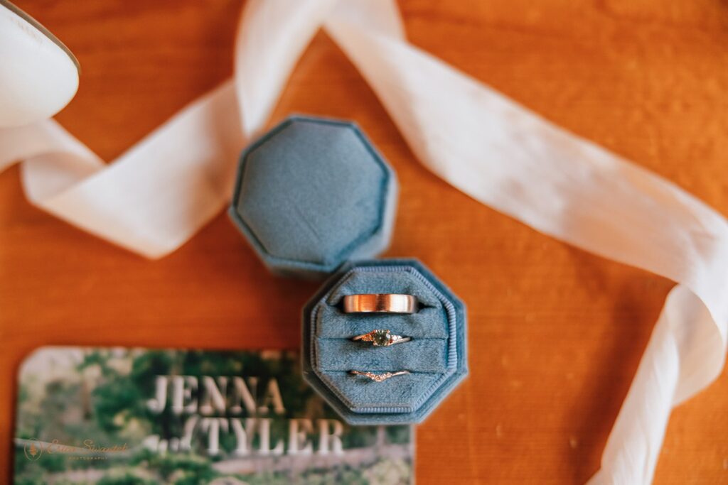 Close-up of wedding rings in blue ring boxes, placed on a wooden surface with a white ribbon and the wedding invitation featuring Jenna and Tyler.