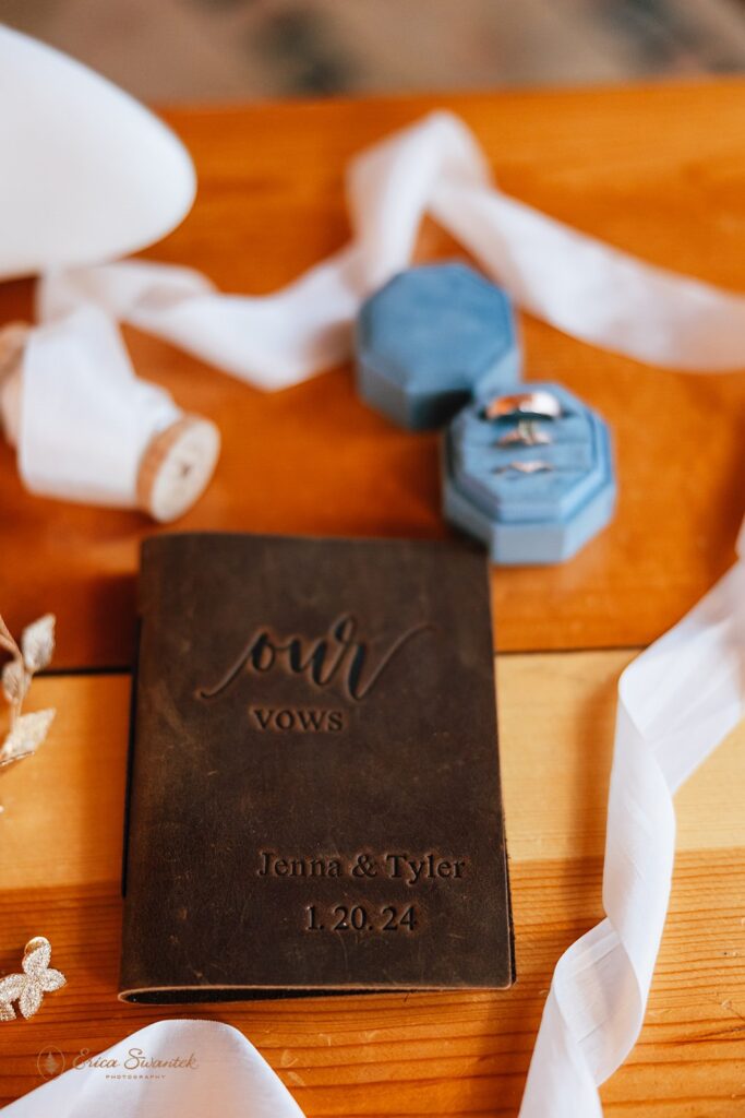 Close-up of a leather-bound vow book engraved with "Our Vows" and the names Jenna & Tyler with the date 1.20.24. Next to it are blue ring boxes, wedding rings, and a white ribbon.