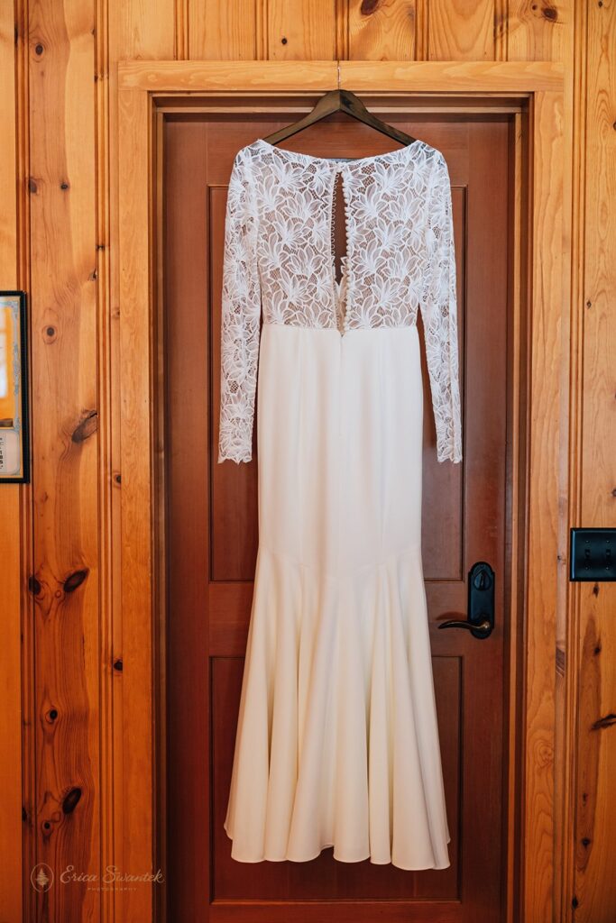 A white wedding dress with a lace top and long sleeves hanging on a wooden door inside a rustic cabin.