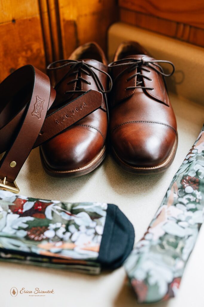 Groom's wedding accessories including brown leather shoes, a matching belt with the wedding date 1.20.2024, floral patterned socks, and a tie displayed on a wooden surface.