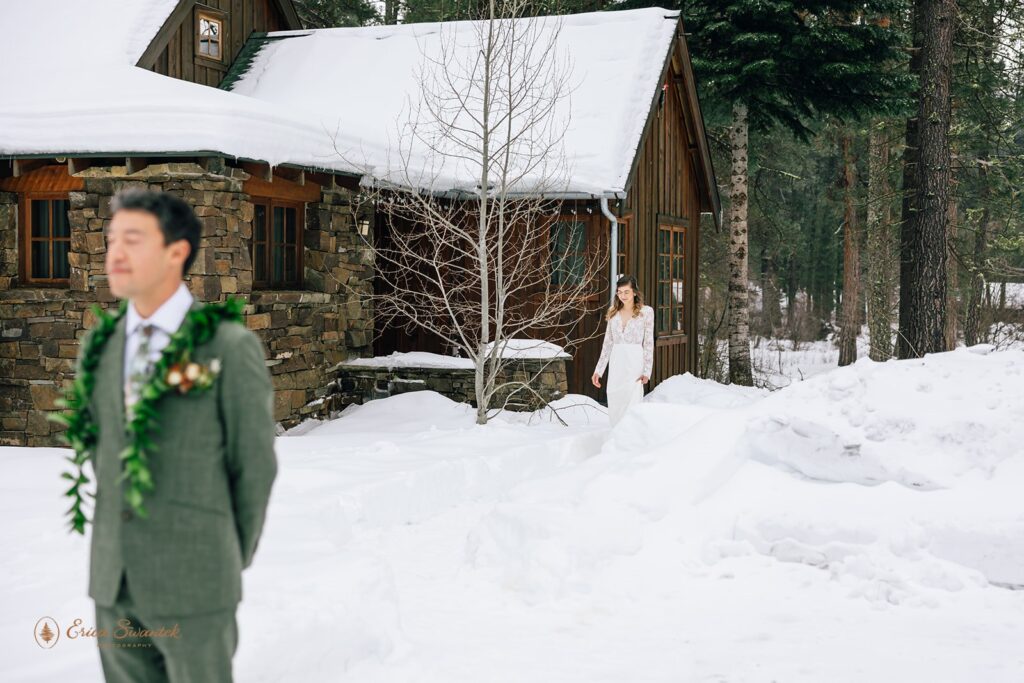 Bride approaches groom for a first look in a snowy setting outside a rustic cabin. Groom is wearing a green suit adorned with a leaf lei, and the bride wears a white lace dress with a plunging neckline.