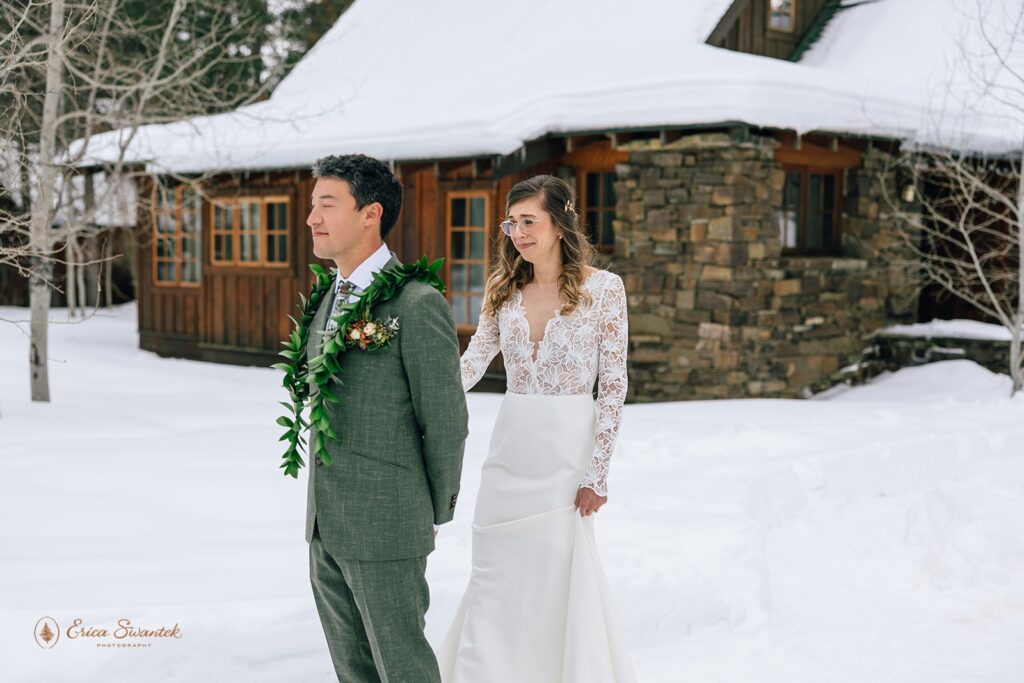 Bride approaches groom for a first look in a snowy setting outside a rustic cabin. Groom is wearing a green suit adorned with a leaf lei, and the bride wears a white lace dress with a plunging neckline.