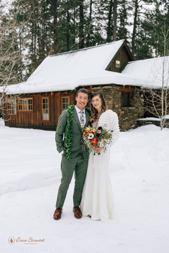 Bride and groom stand hand in hand in front of a snow-covered cabin. The groom wears a green suit with a leaf lei, while the bride wears a white lace dress and holds a colorful bouquet.