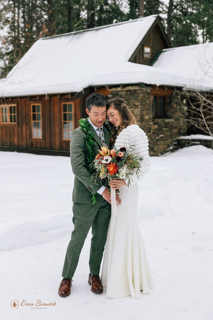 Bride and groom stand hand in hand in front of a snow-covered cabin. The groom wears a green suit with a leaf lei, while the bride wears a white lace dress and holds a colorful bouquet.