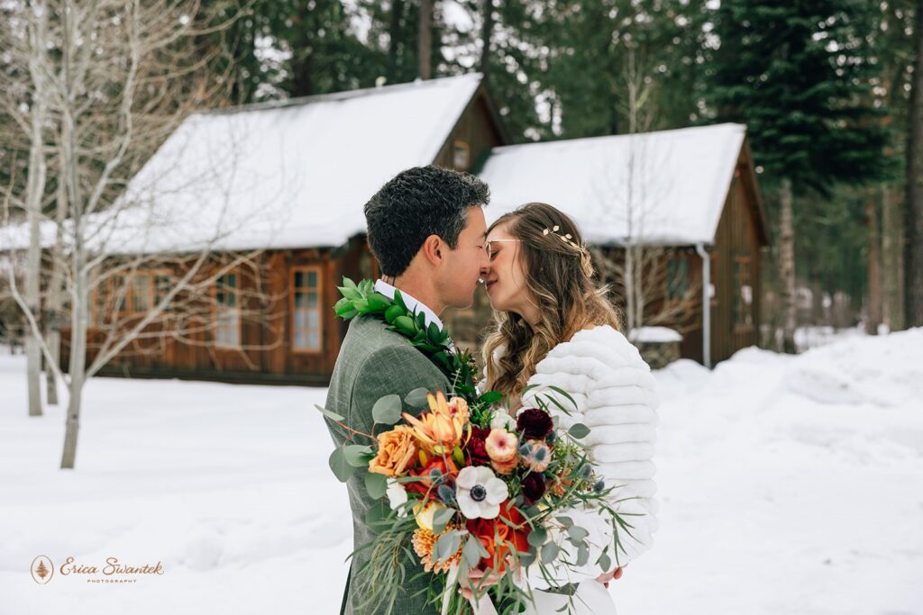 Bride and groom share an intimate moment forehead to forehead in the snow. The bride holds a vibrant bouquet, and the groom wears a green suit with a leaf lei.