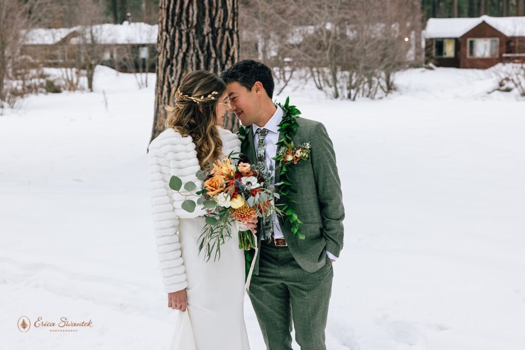 Bride and groom share an intimate moment forehead to forehead in the snow. The bride holds a vibrant bouquet, and the groom wears a green suit with a leaf lei.