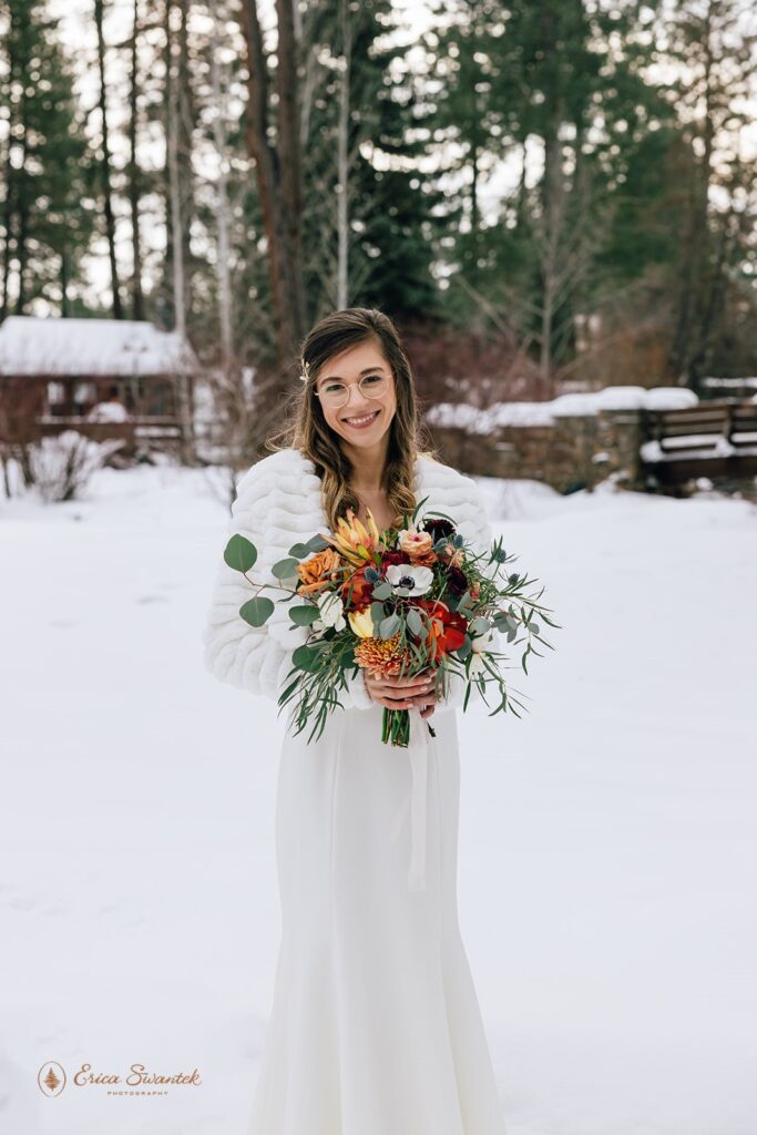 Beautiful winter wedding bride outdoor portrait surrounded by snow. She wears a white lace dress, a white fluffy jacket and holds a colorful bouquet.