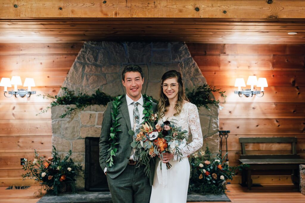 Bride and groom with pinecones and greenery inside a cozy cabin with a stone fireplace in the background.