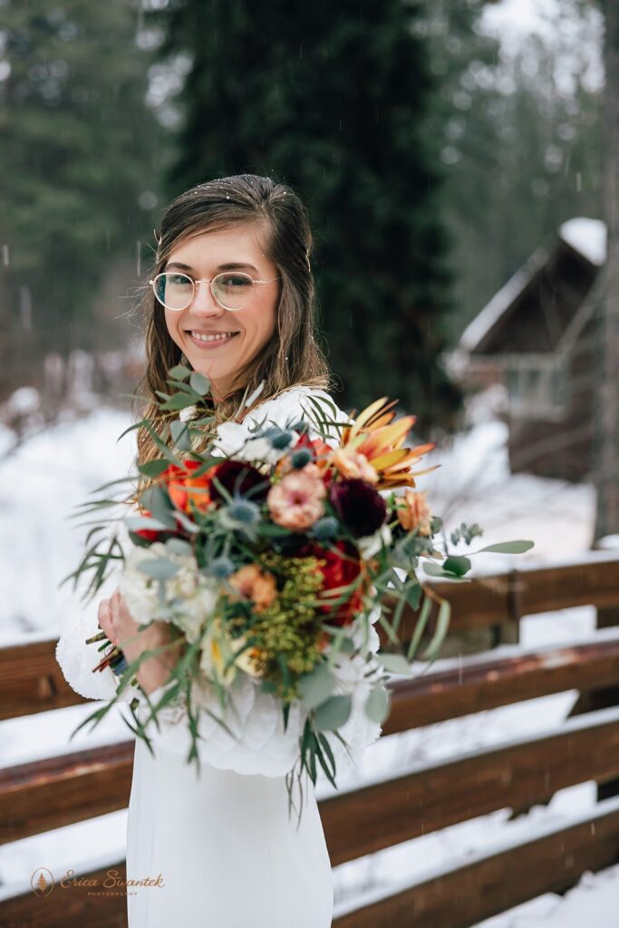 Beautiful winter wedding bride outdoor portrait surrounded by snow. She wears a white lace dress, a white fluffy jacket and holds a colorful bouquet.