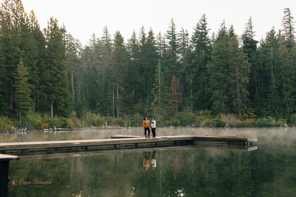 A LGBTQ+ couple stands holding hands on the boat dock of Suttle Lake as the morning fog lifts during their adventure engagement session.
