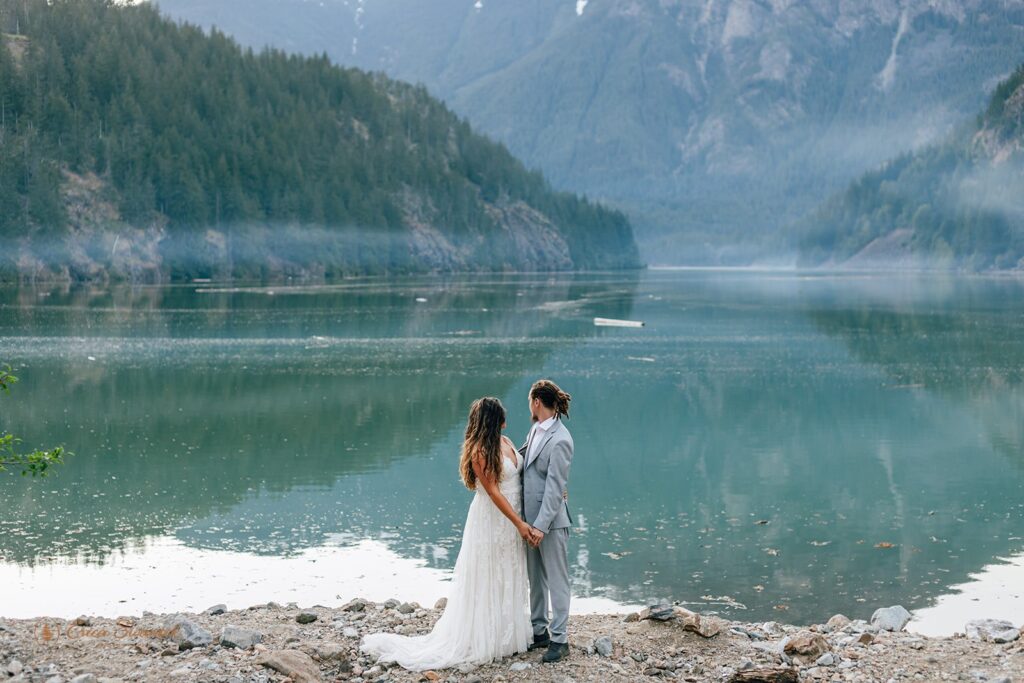 bride and groom standing at a crystal clear lake shore with mountains in the backdrop