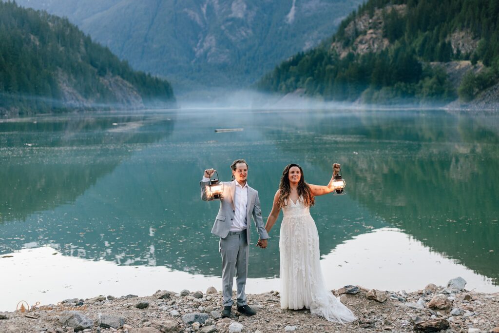 bride and groom holding lanterns standing at a lake shore