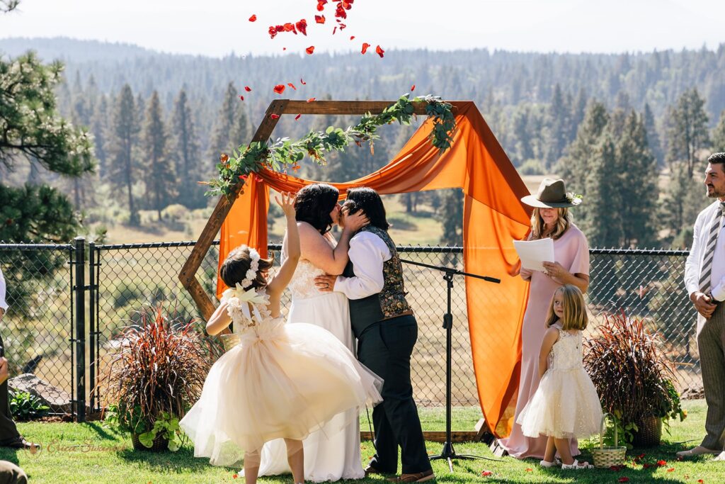 LGBTQ+ couple kissing at their wedding as their daughter tosses rose petals in the air during their intimate Bend Oregon elopement. 