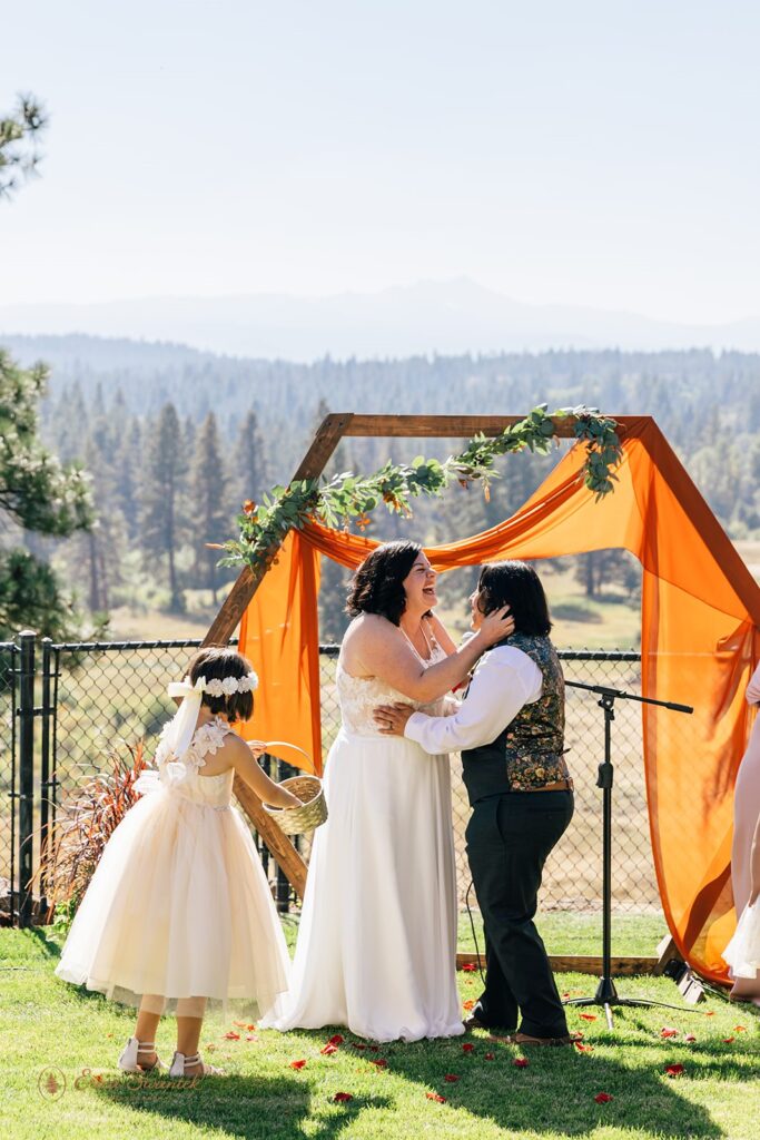 Two brides embrace as their daughter throws rose petals during their intimate Oregon wedding at Suttle Lake in Oregon.