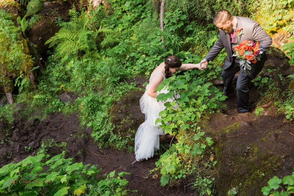 A groom helps a bride up a slippery muddy trail in her wedding gown.