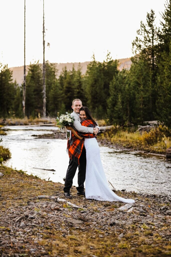 A bride and groom are wrapped in a red checkered blanket standing by a creek near Sparks Lake, Oregon. 