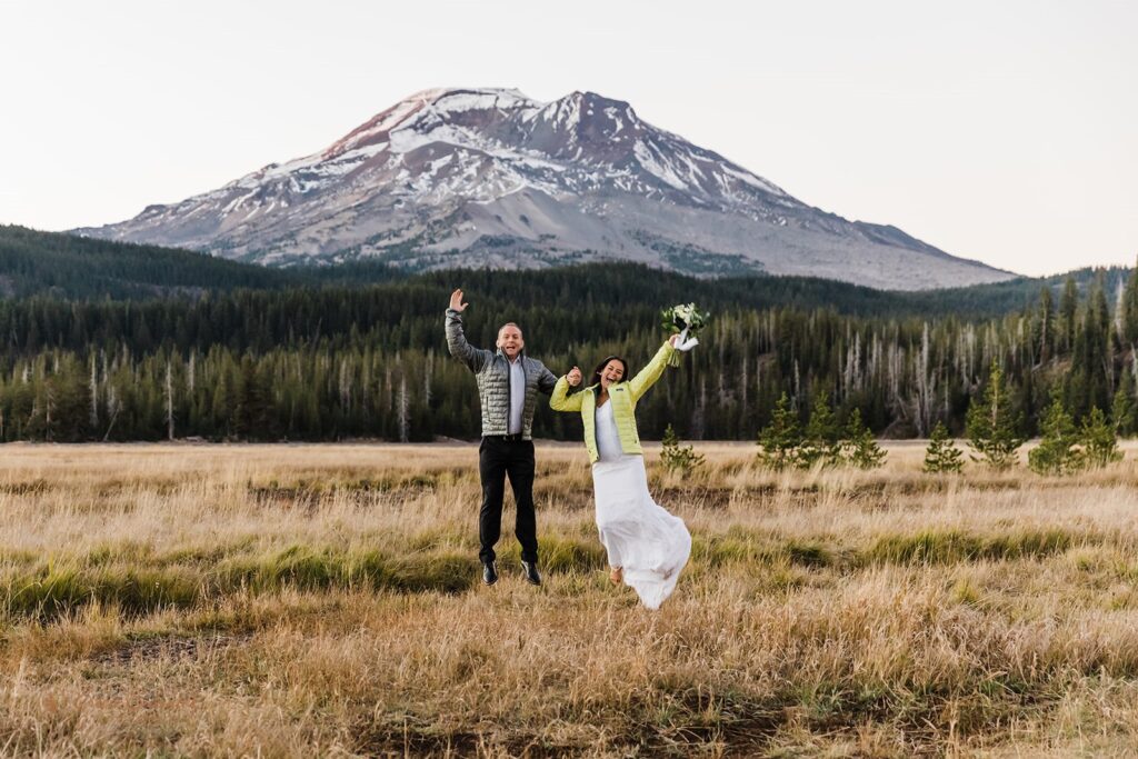 Bride and groom jump up in front of a mountain wearing their Patagonia puffy jackets.