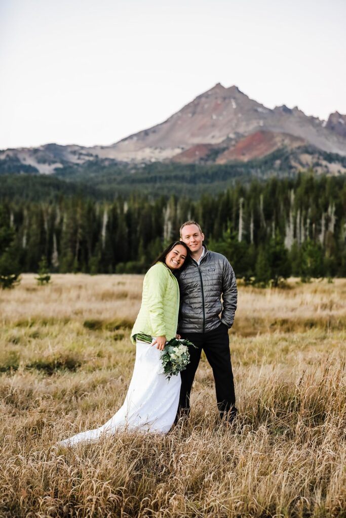 A bride and groom wear down puffy jackets with mountains in the background.