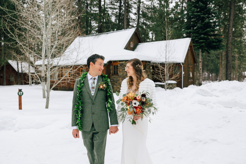 A groom wearing a green suit and lea holds his bride's hand while she carries the bouquet in her other hand as they walk through the snow at Lake Creek Lodge in Camp Sherman, Oregon. 