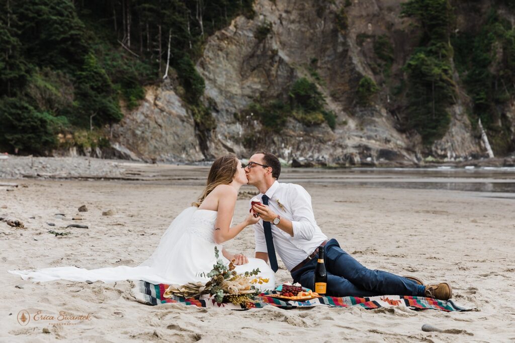 A couple kisses holding cupcakes while sitting on a blanket at the beach with a picnic.