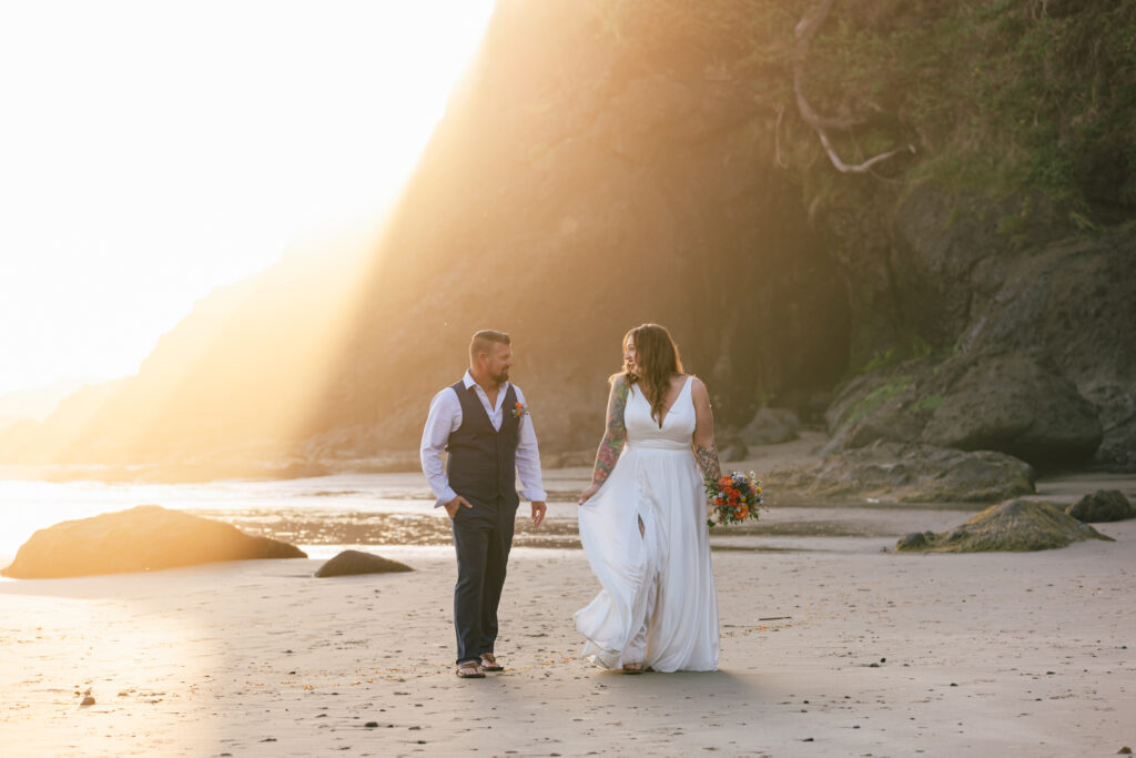 Bride and groom walk hand in hand on the Oregon Coast beach with sun rays behind them.