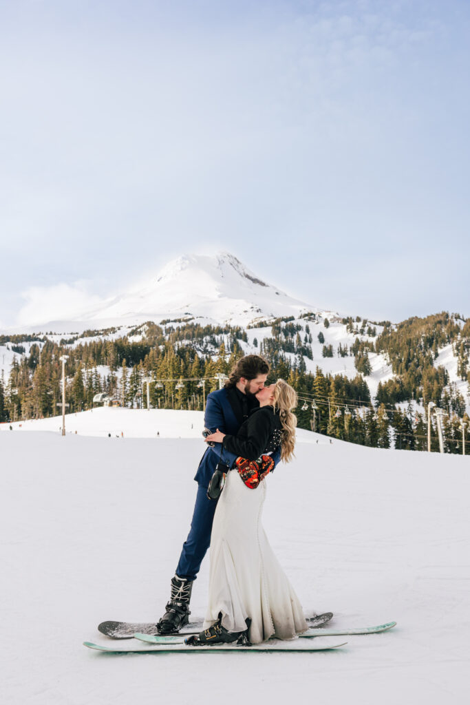 A groom on a snowboard embraces and kisses his bride on skis at Mt. Hood is off in the distance at their Oregon ski resort elopement. 