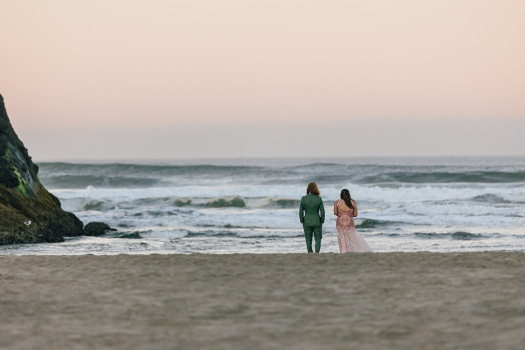 A couple stands on the shore of the Pacific Ocean as the sunrise lights up the sky. 