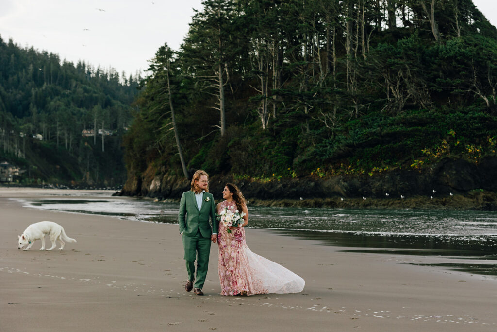 A bride and groom walk along the beach as their dog follows behind.