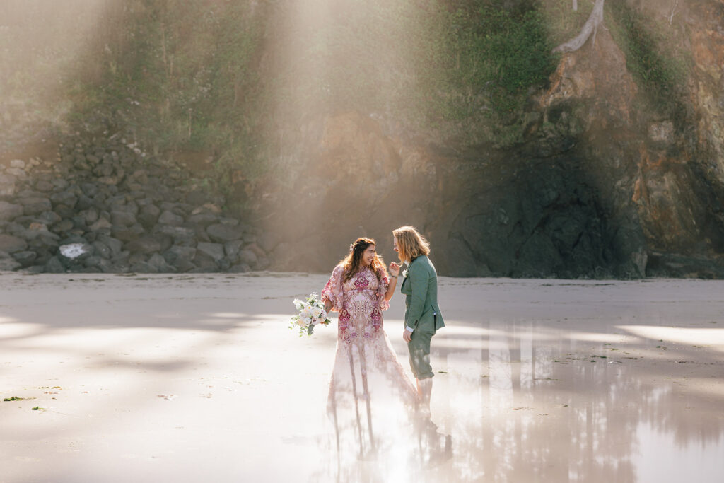 A gorgeous bride in a pink boho wedding dress dances with her groom wearing a green suite in the sunlight on the beach. 
