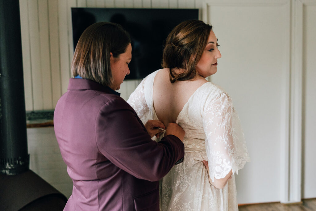 A bride in a mauve suit helps her bride zip her boho wedding dress.