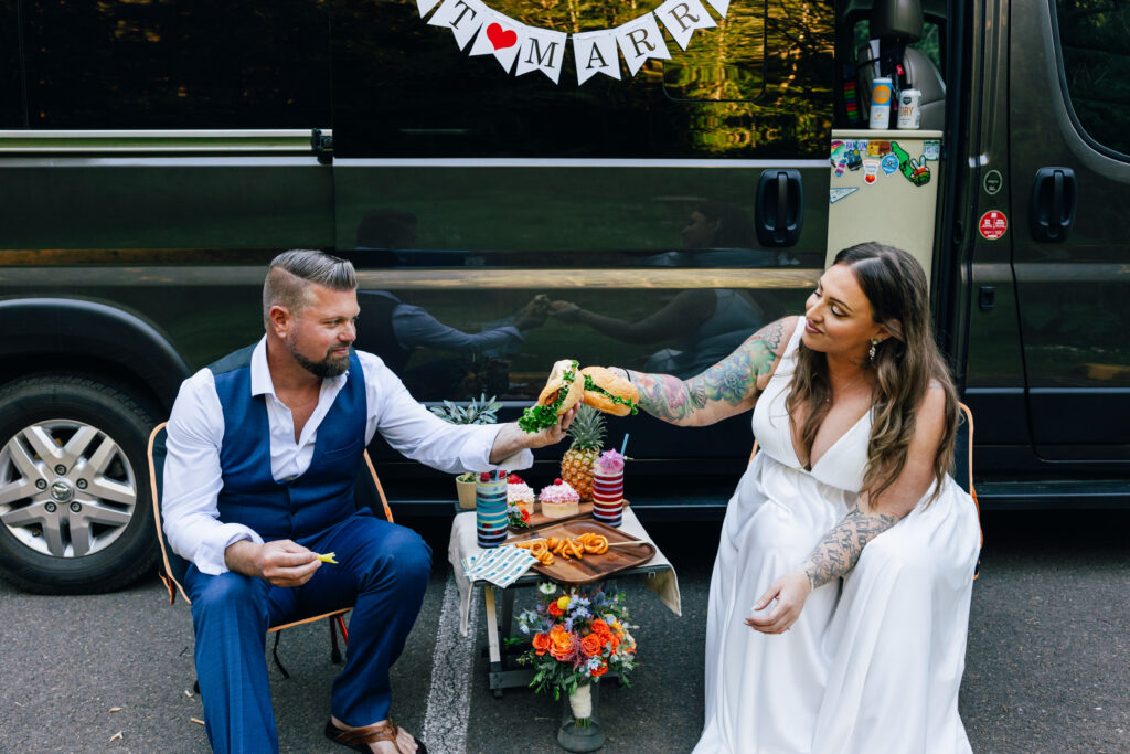 A bride and groom sit in front of their adventure van with food they made for their elopement day. 