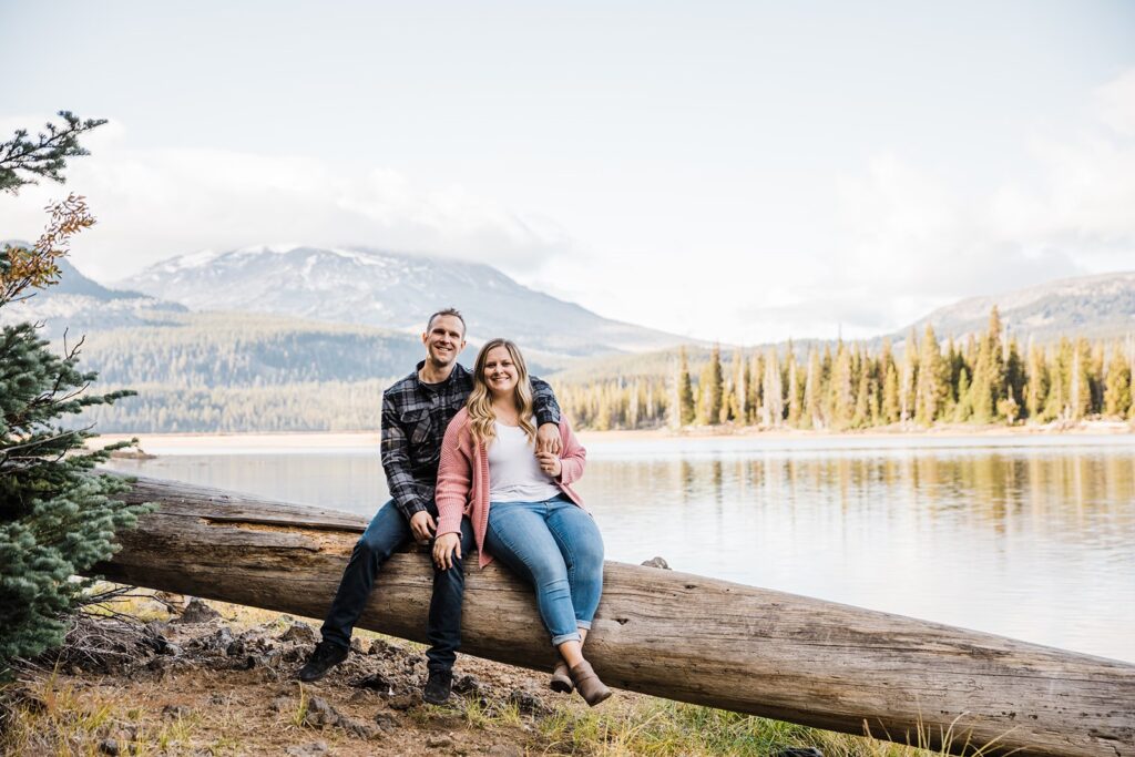 cuddly couple sitting on a large log with beautiful lake and mountain views in the backdrop