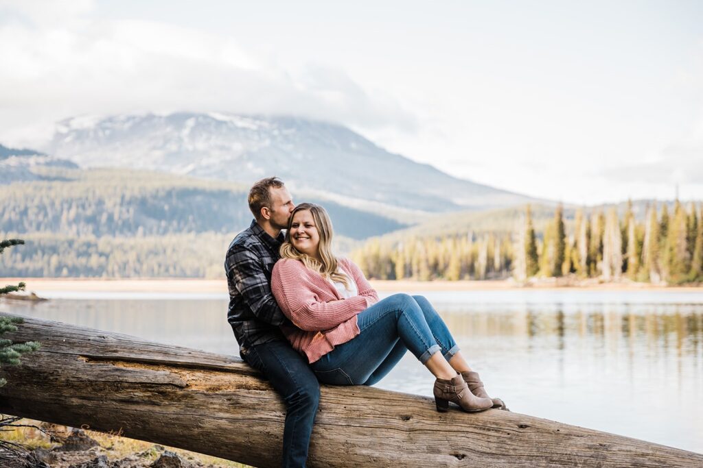 cuddly couple sitting on a large log with beautiful lake and mountain views in the backdrop