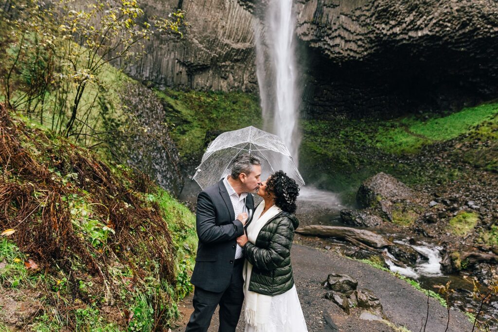 bride and groom under clear umbrellas during their rainy PNW elopement with waterfall backdrop