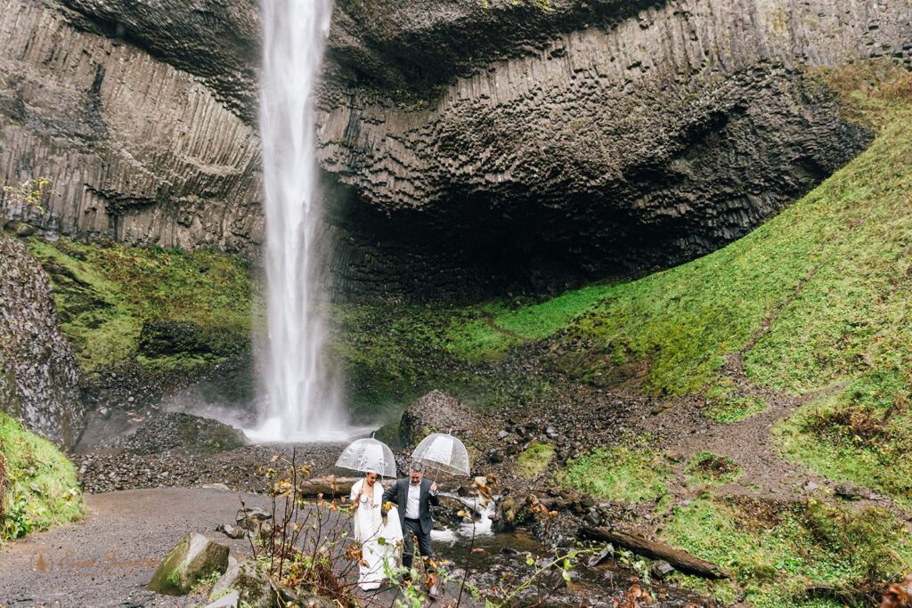 bride and groom under clear umbrellas during their rainy PNW elopement with waterfall backdrop