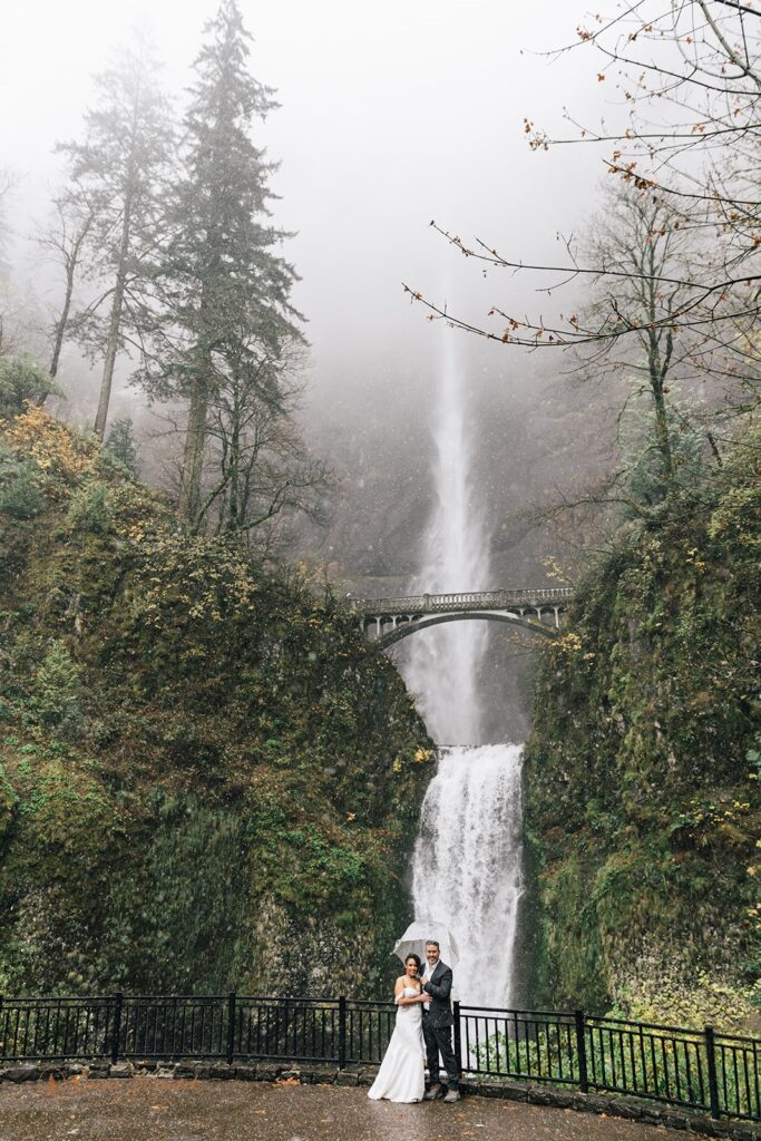 bride and groom under clear umbrellas during their rainy PNW elopement with waterfall backdrop