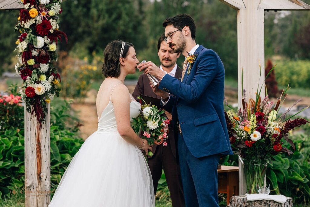 The groom offers the bride a drink from a kiddush cup during the wedding ceremony under a floral-adorned wooden arch