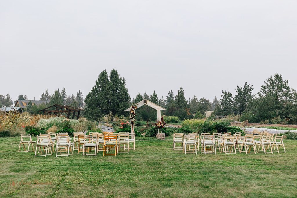 Outdoor wedding ceremony setup with wooden chairs arranged on green grass in front of a simple wooden arch adorned with flowers at Rainshadow Organics Farm wedding day