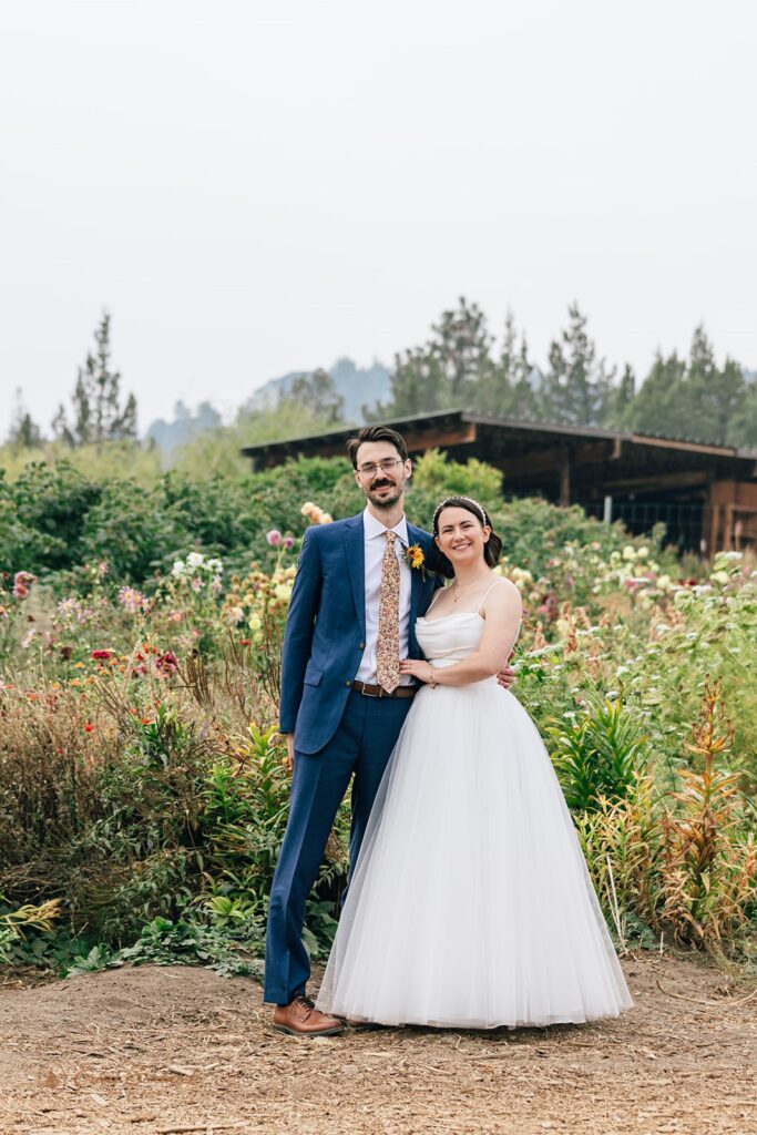 The bride and groom standing close together in front of a vibrant garden of flowers, smiling at the camera during their Rainshadow Organics Farm wedding