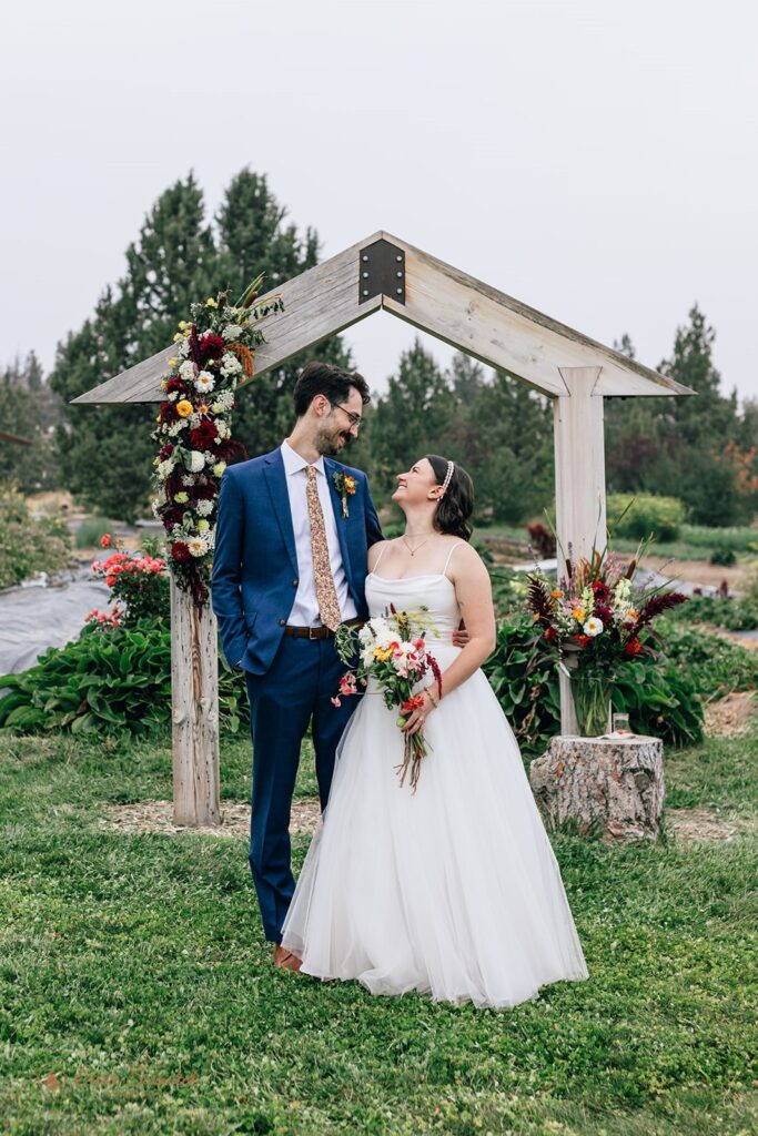 The bride and groom sharing an intimate moment under a wooden wedding arch decorated with flowers, surrounded by greenery at the Rainshadow Organics Farm