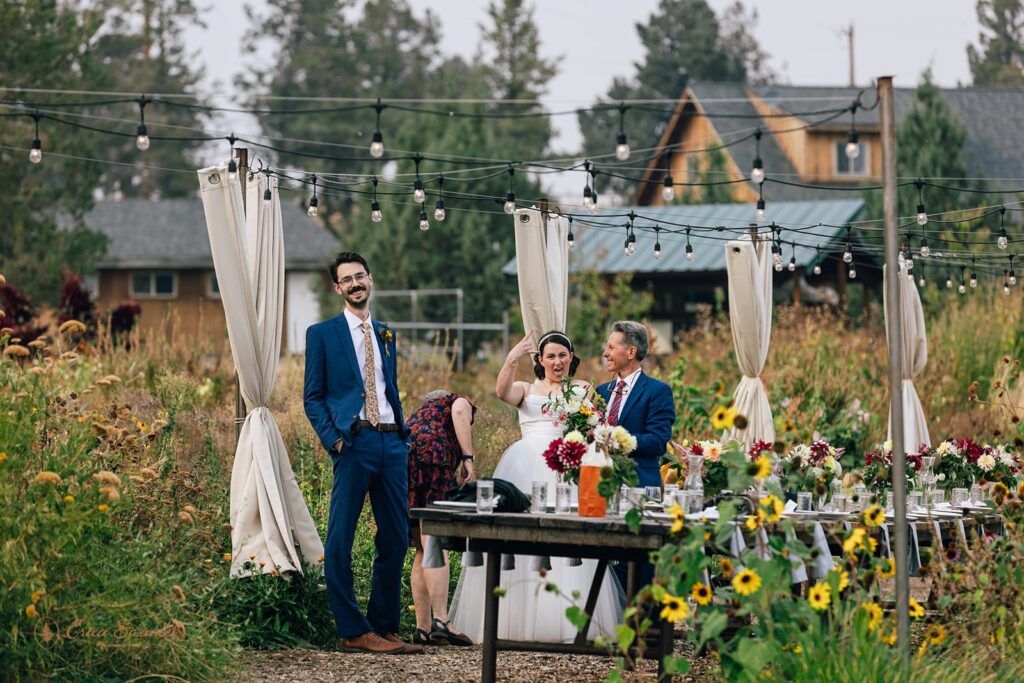 A wedding party enjoying the reception in a garden setting with string lights and rustic decor, featuring the groom in a blue suit and the bride in a white dress