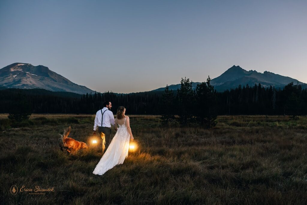 moody pnw elopement photo with the couple walking through the field with lanterns in hands with their dog running next to them