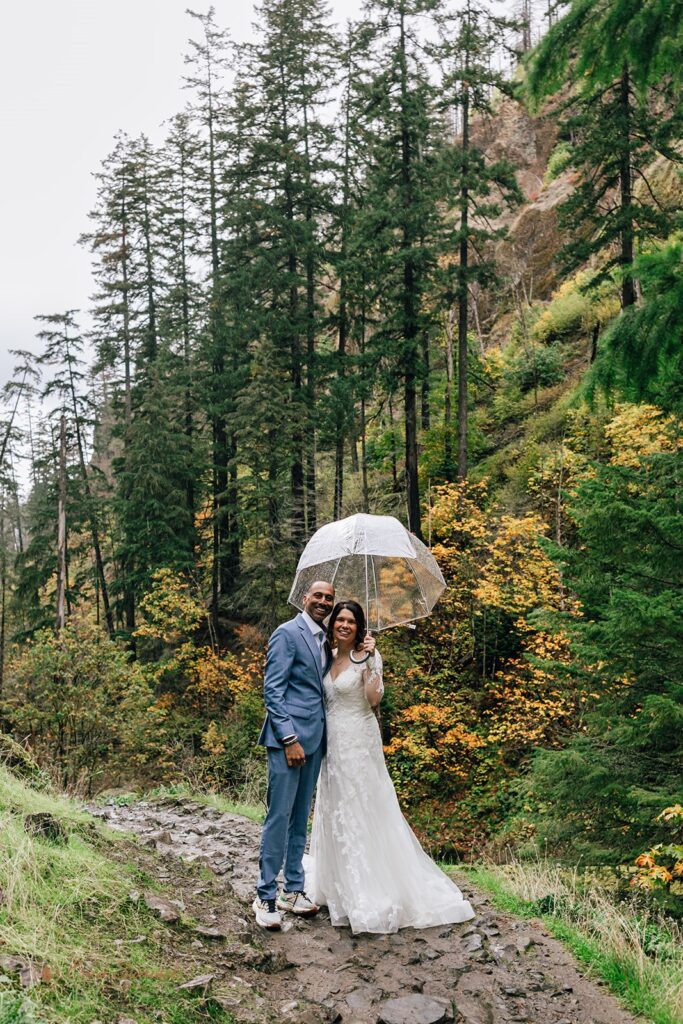 rainy waterfall elopement day in oregon, couple walking under a clear umbrella